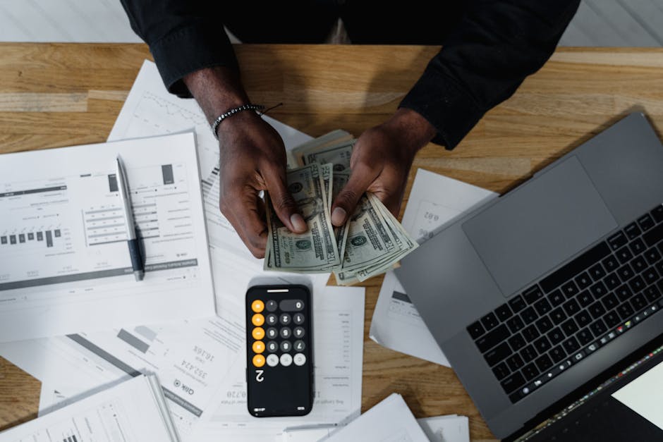 Person counting US dollars, using a calculator and laptop, with financial documents on a wooden desk